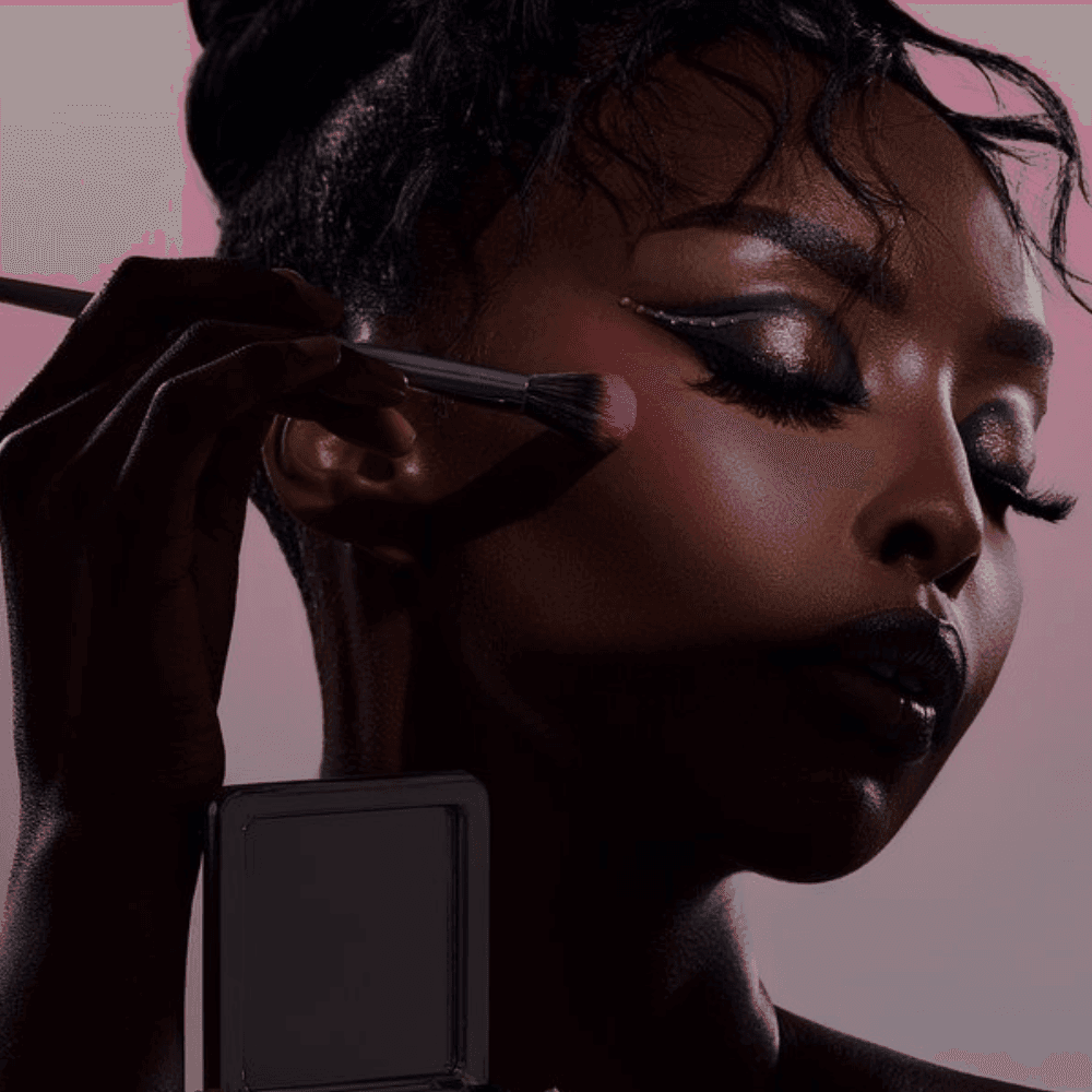 Person applying blush with a makeup brush, showcasing dramatic eye makeup and dark lipstick.